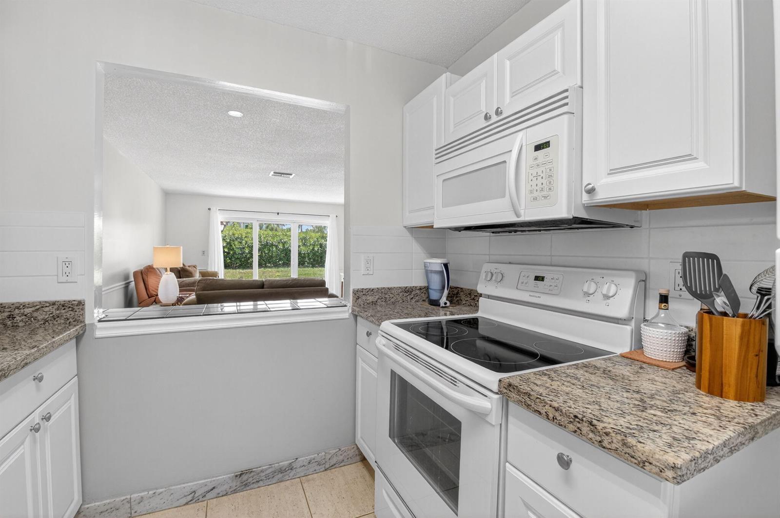 A kitchen with white cabinets, granite countertops, and a pass-through window looking into a living room — 13984 Vía Flora # B, Delray Beach FL 33484, DELRAY BEACH, FL