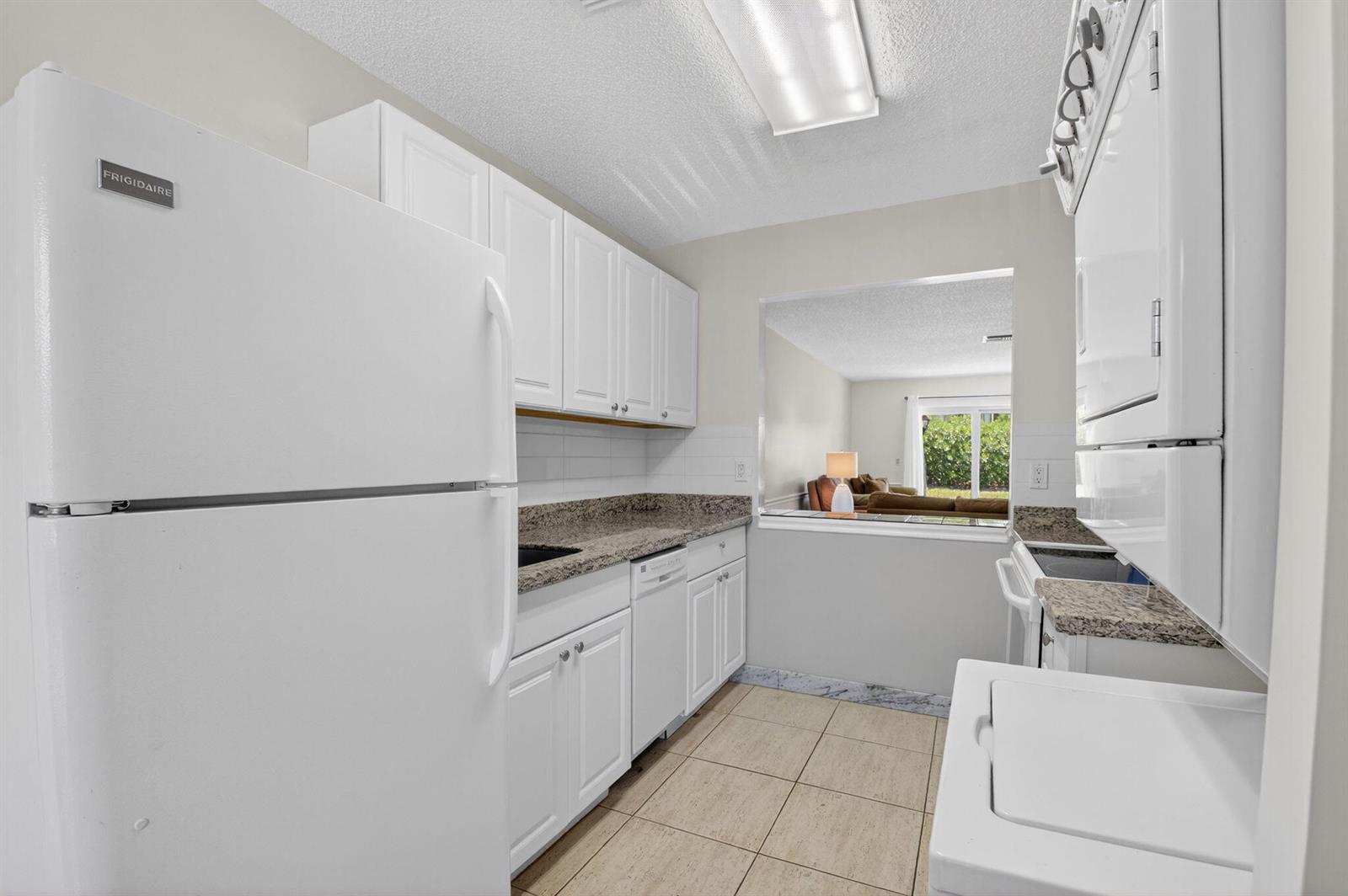 A white kitchen features granite countertops, white cabinetry, and a pass-through window to the living area — 13984 Vía Flora # B, Delray Beach FL 33484, DELRAY BEACH, FL