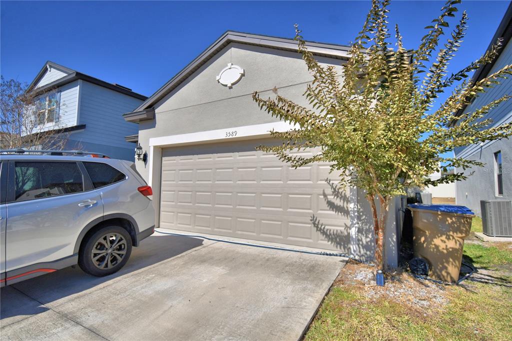 A beige house exterior features a two-car garage, a concrete driveway, and a small tree — 3589 ROCK CREEK LANE, KISSIMMEE, FL
