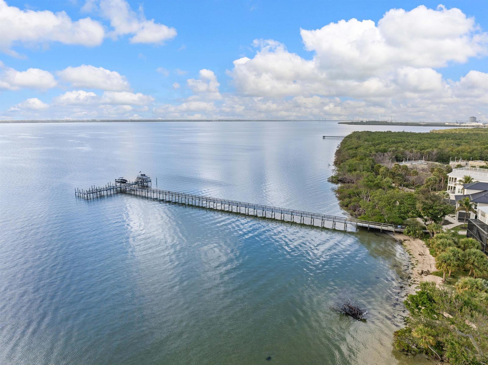 An aerial view shows a long wooden pier extending into a calm bay near trees — 7671 Pelican Pointe Drive, Jensen Beach FL 34957, JENSEN BEACH, FL