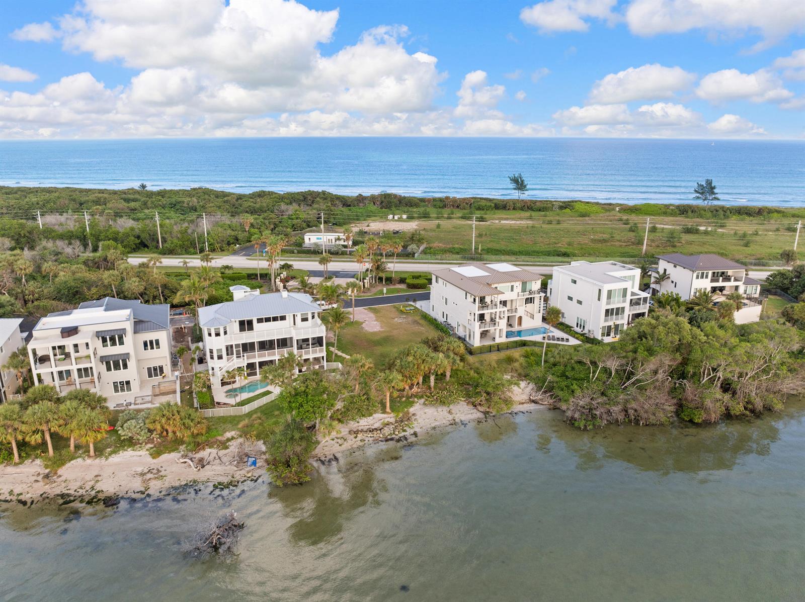 An aerial view shows several multi-story houses situated along a waterfront with a nearby road — 7671 Pelican Pointe Drive, Jensen Beach FL 34957, JENSEN BEACH, FL