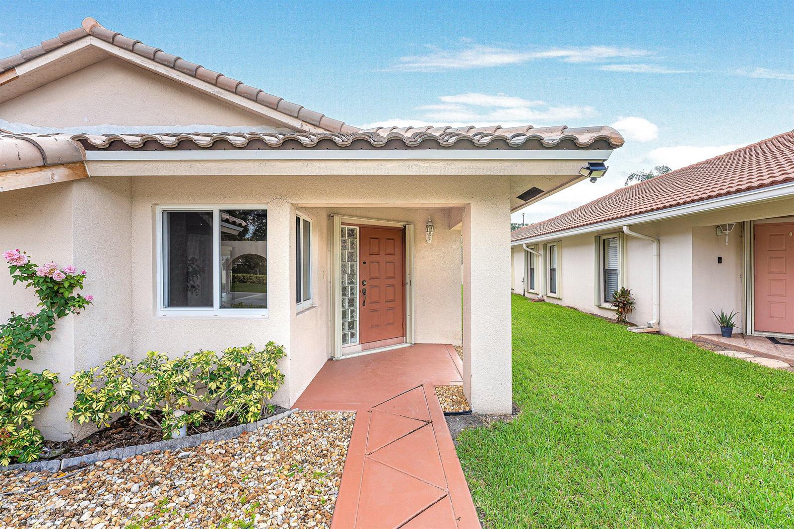 A tan stucco house exterior features a covered entryway with a brown door and walkway — 6290 Sweet Maple Lane, Boca Raton FL 33433, BOCA RATON, FL
