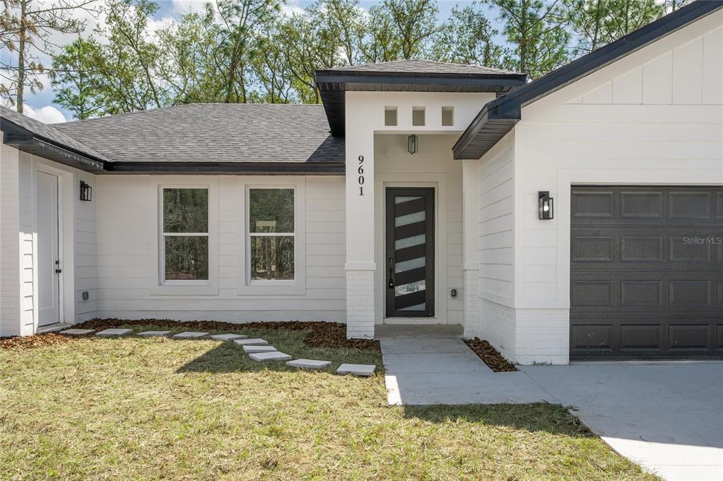 The exterior of a white house features a dark front door, garage, and shingled roof — 9609 N VAUGHN DRIVE, CITRUS SPRINGS, FL