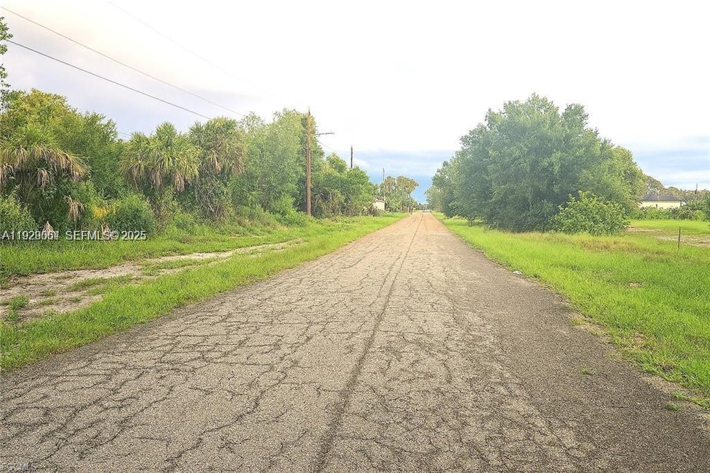 A narrow, cracked asphalt road stretches through a rural area with trees and grassy fields — 613 Grant Blvd, Lehigh Acres FL 33974, LEHIGH ACRES, FL