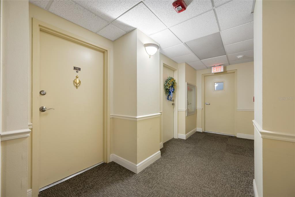 A hallway with beige walls, carpeted flooring, two doors, and an exit sign — 1621 GULF BOULEVARD UNIT 501, CLEARWATER BEACH, FL