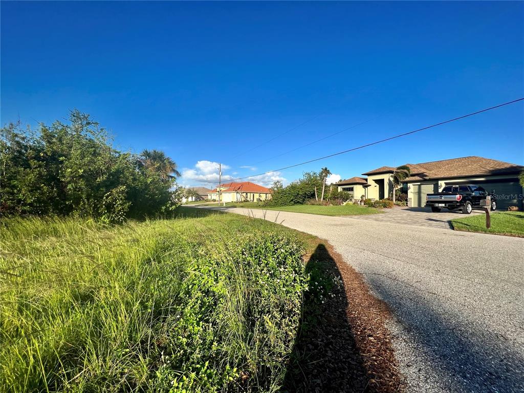 A paved road curves past a grassy lot toward houses under a blue sky — 13468 RAYBURN LANE, PORT CHARLOTTE, FL