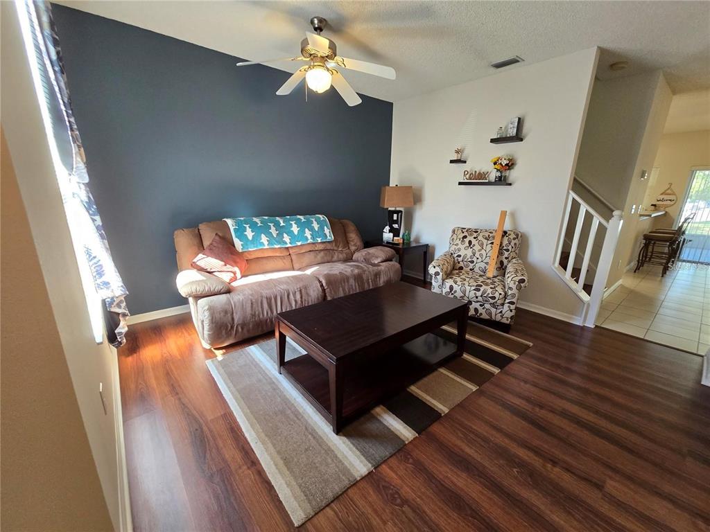 A living room features a dark accent wall, brown sofa, patterned armchair, and wood flooring — 30119 WELLESLEY WAY, WESLEY CHAPEL, FL