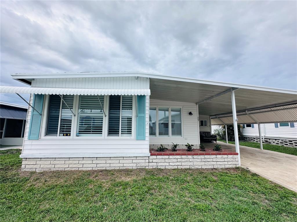 A white mobile home features a carport, brick skirting, and light blue window shutters — 1100 BELCHER ROAD S UNIT 727, LARGO, FL