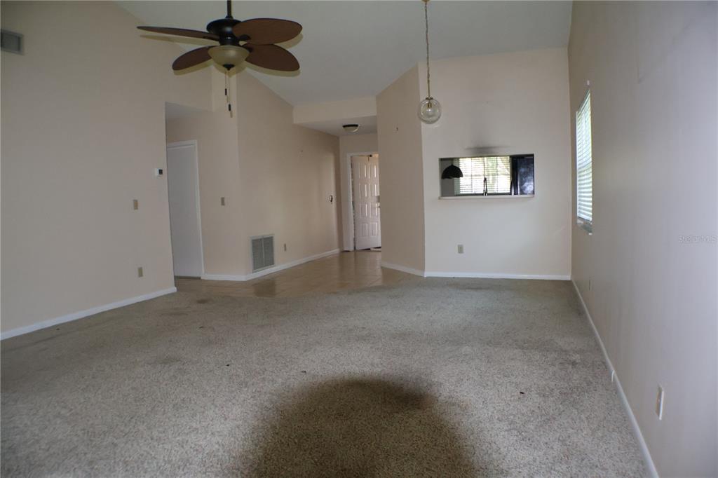 An empty living room with beige carpet, a ceiling fan, and a kitchen pass-through window — 11806 WHITE ASH DRIVE, NEW PORT RICHEY, FL