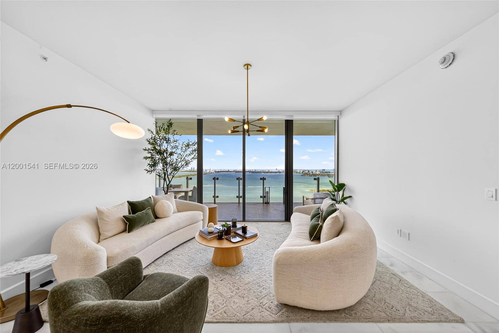 A modern living room features two curved sofas, a coffee table, and floor-to-ceiling water views — 700 NE 26 Ter # 3602, Miami FL 33137, MIAMI, FL