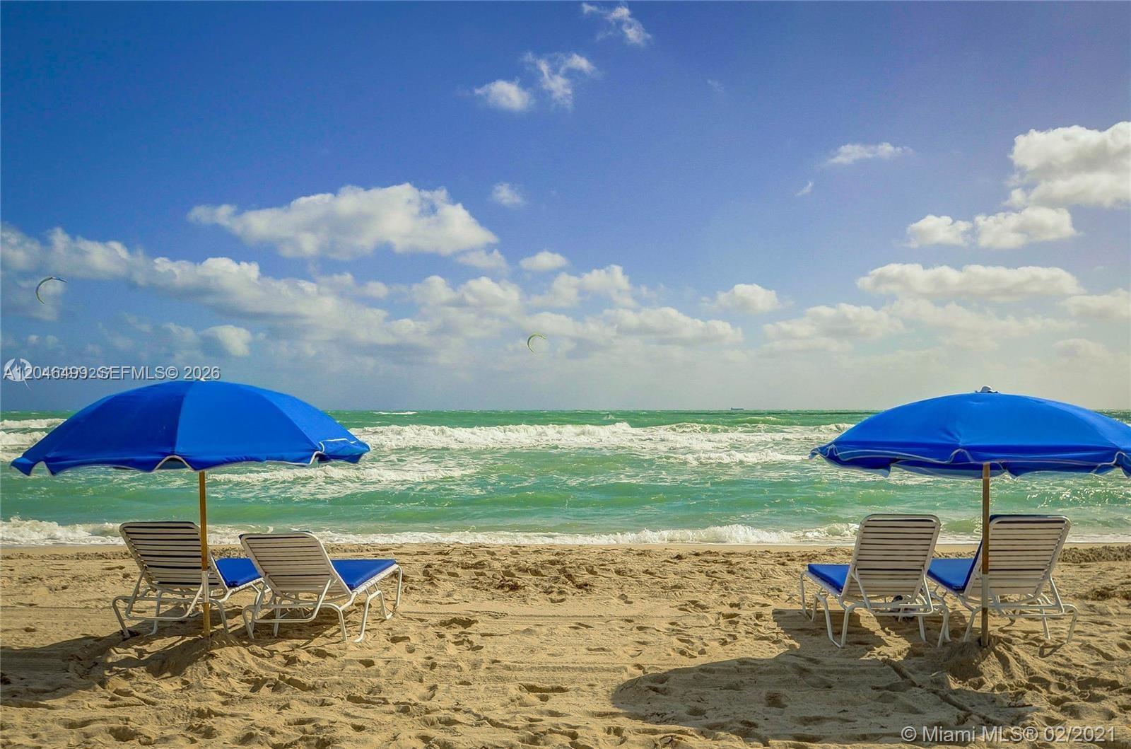 Two blue beach umbrellas shade four lounge chairs on a sandy beach by the ocean — 6301 Collins Ave # 807, Miami Beach FL 33141, MIAMI BEACH, FL