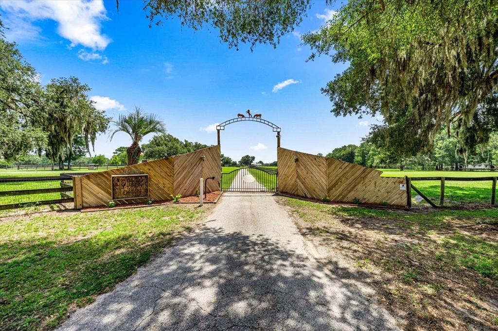 A paved driveway leads to a gated entrance with wooden fencing and an arched sign — 5065 NW 150TH AVENUE, MORRISTON, FL