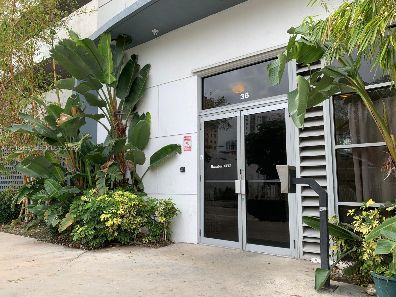 The exterior of a modern multi-story building features a glass entrance door and lush landscaping — 36 NW 6th Ave # 1108, Miami FL 33128, MIAMI, FL