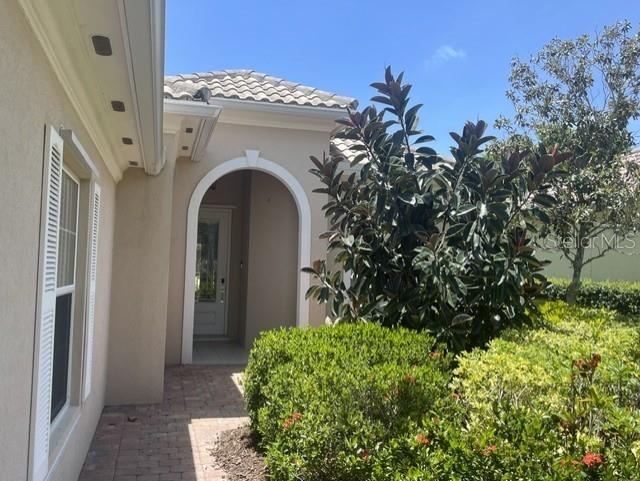 A beige house exterior features an arched entryway, a brick walkway, and a large shrub — 11818 FIORE LANE, SARASOTA, FL