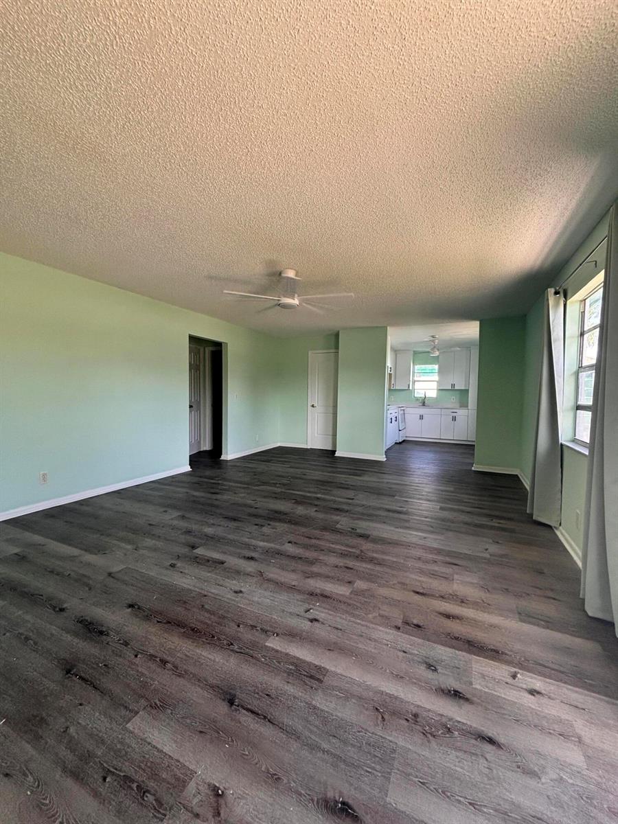 A spacious living room with light green walls and dark wood-look flooring leads to a kitchen — 1328 W 6th Street, Riviera Beach FL 33404, RIVIERA BEACH, FL