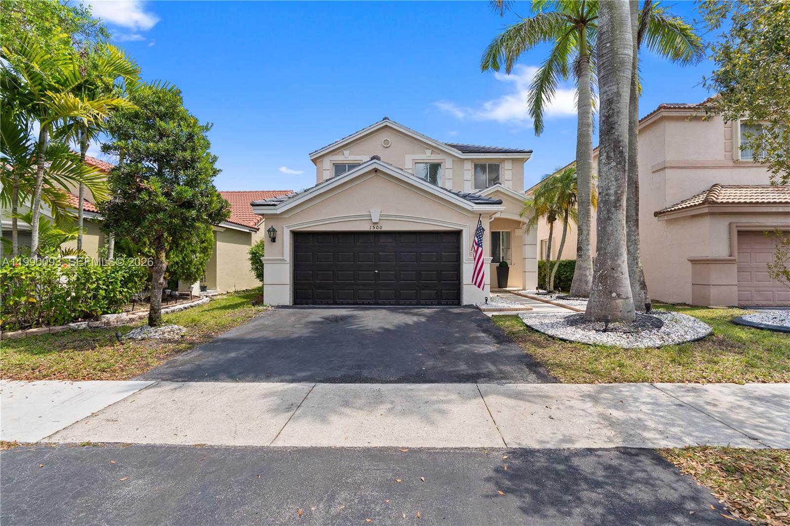 A two-story beige house features a dark garage door, a palm tree, and an American flag — 1500 Canary Island Dr, Weston FL 33327, WESTON, FL