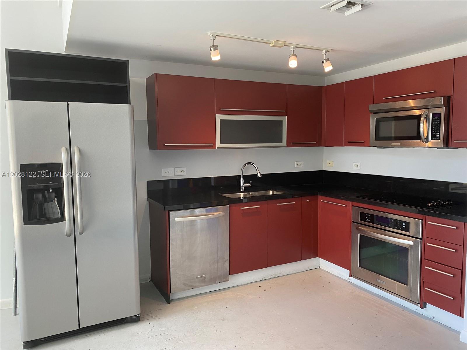 A kitchen features red cabinets, stainless steel appliances, and black countertops against a white wall — 253 NE 2nd St # 621, Miami FL 33132, MIAMI, FL