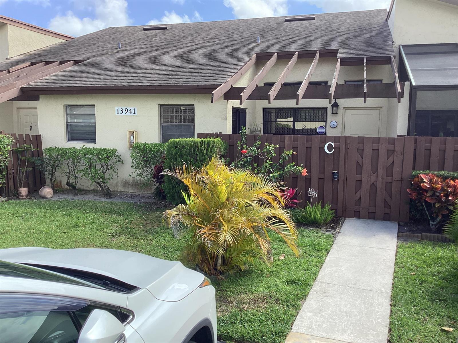 A single-story residential unit features a brown wooden fence, a walkway, and a palm plant — 13941 Nesting Way # C, Delray Beach FL 33484, DELRAY BEACH, FL