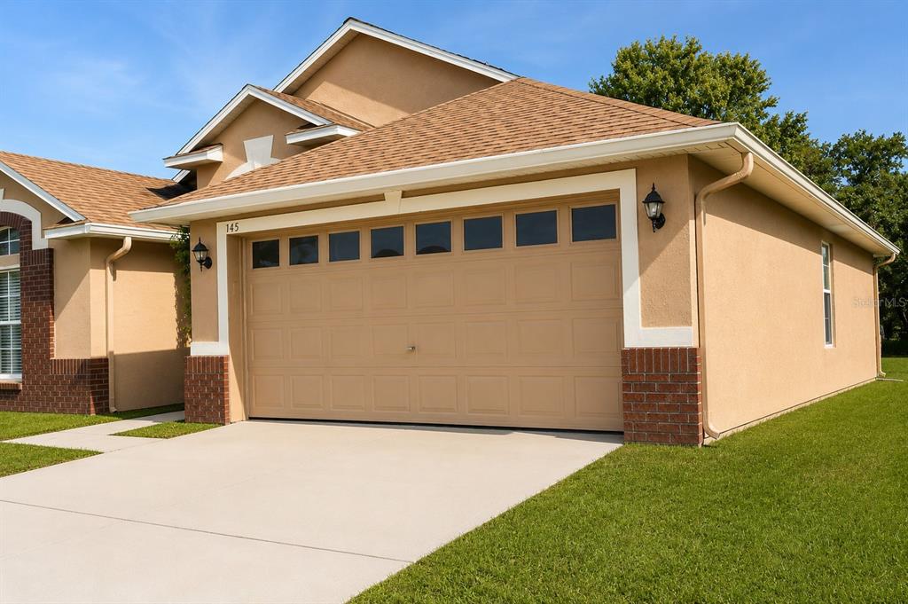 A tan stucco house features a two-car garage, a brown shingled roof, and brick accents — 145 THORNBURY DRIVE, KISSIMMEE, FL