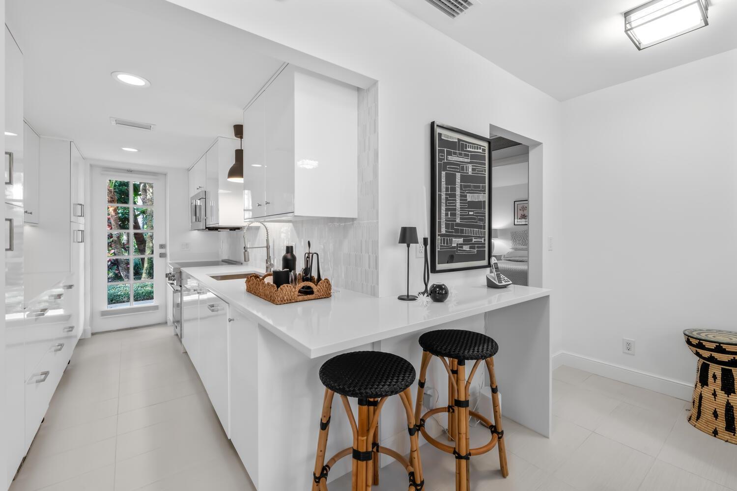 A modern white kitchen features a breakfast bar with two stools and a glass door — 134 Peruvian Avenue # 104, Palm Beach FL 33480, PALM BEACH, FL