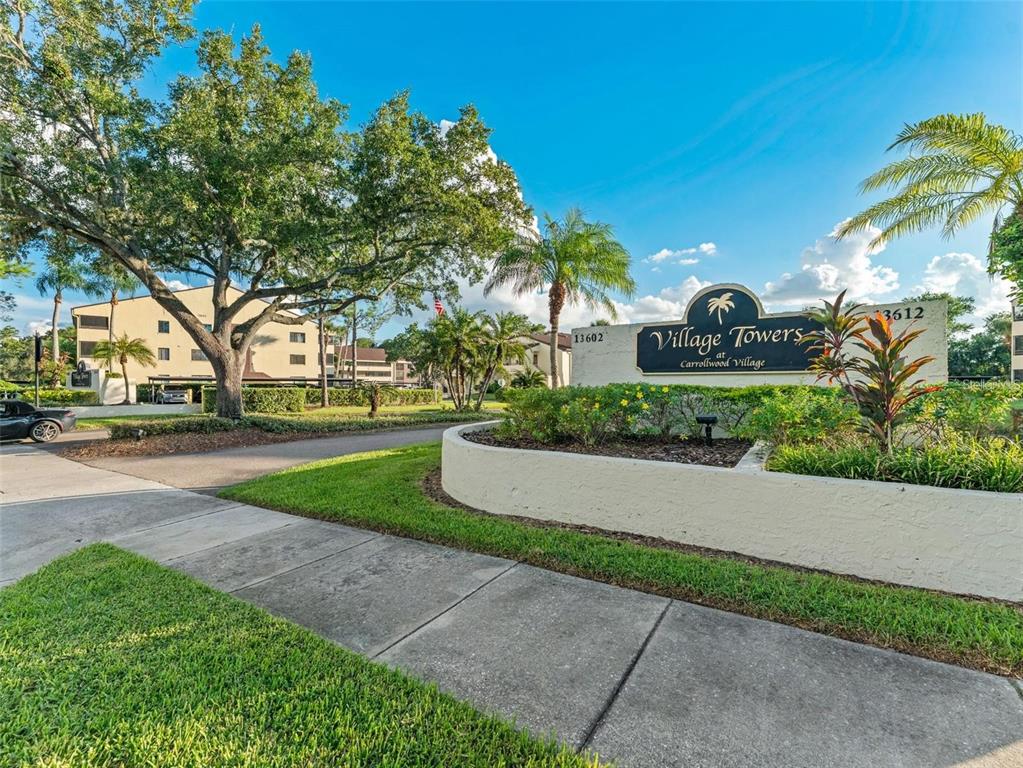 A sign for Village Towers at Carrollwood Village stands near a sidewalk and large trees — 13608 S VILLAGE DRIVE UNIT 7204, TAMPA, FL