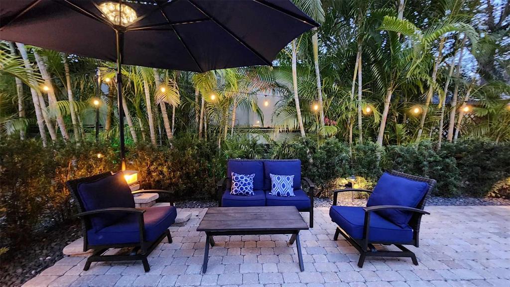 A patio features a dark umbrella, a loveseat, two chairs, and string lights among palm trees — 140 APPALOOSA TRAIL, RIVER RANCH, FL