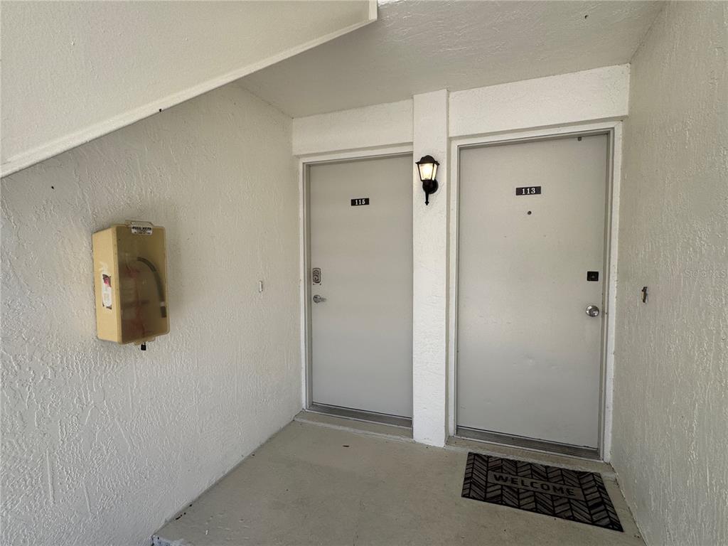 Two apartment doors numbered 113 and 115 are located in a white stucco outdoor entryway — 115 WINDWARD PLACE UNIT 15, OLDSMAR, FL