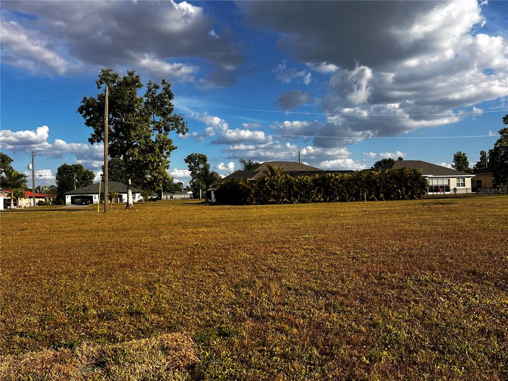 A large, flat, grassy vacant lot is surrounded by residential houses and trees under a cloudy sky — 25458 ISLAS DRIVE, PUNTA GORDA, FL