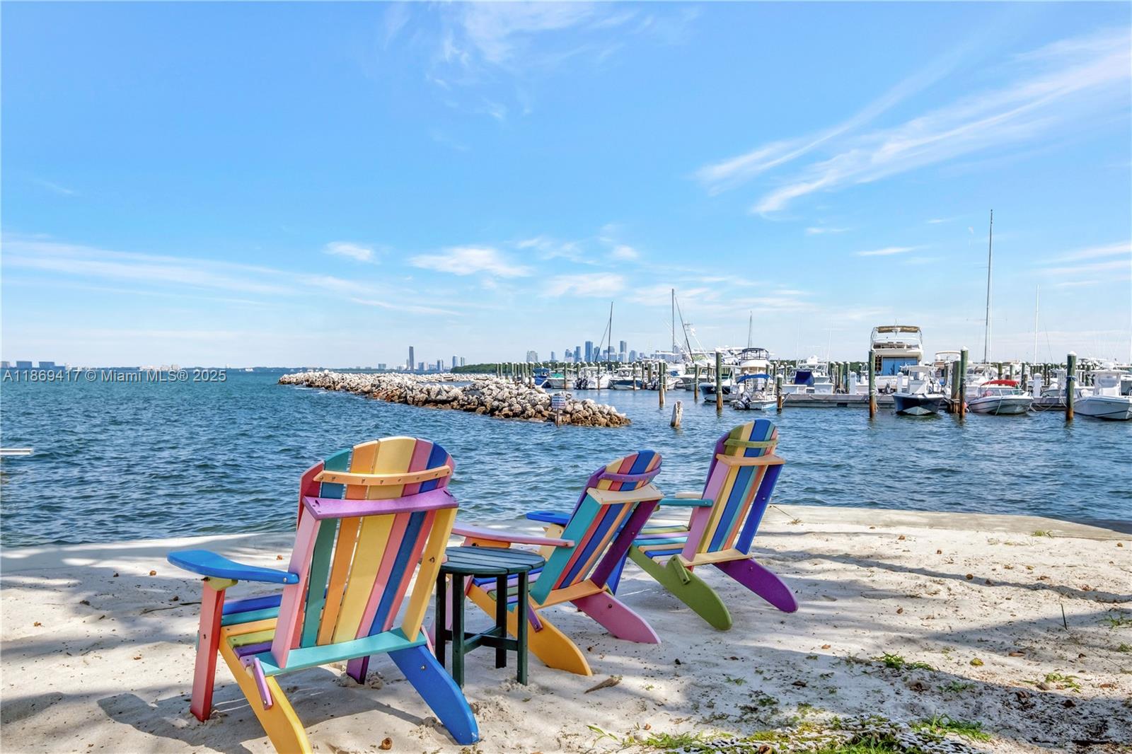Three colorful Adirondack chairs sit on a sandy beach overlooking a marina and city skyline — 200 Harbor Dr, Key Biscayne FL 33149, KEY BISCAYNE, FL