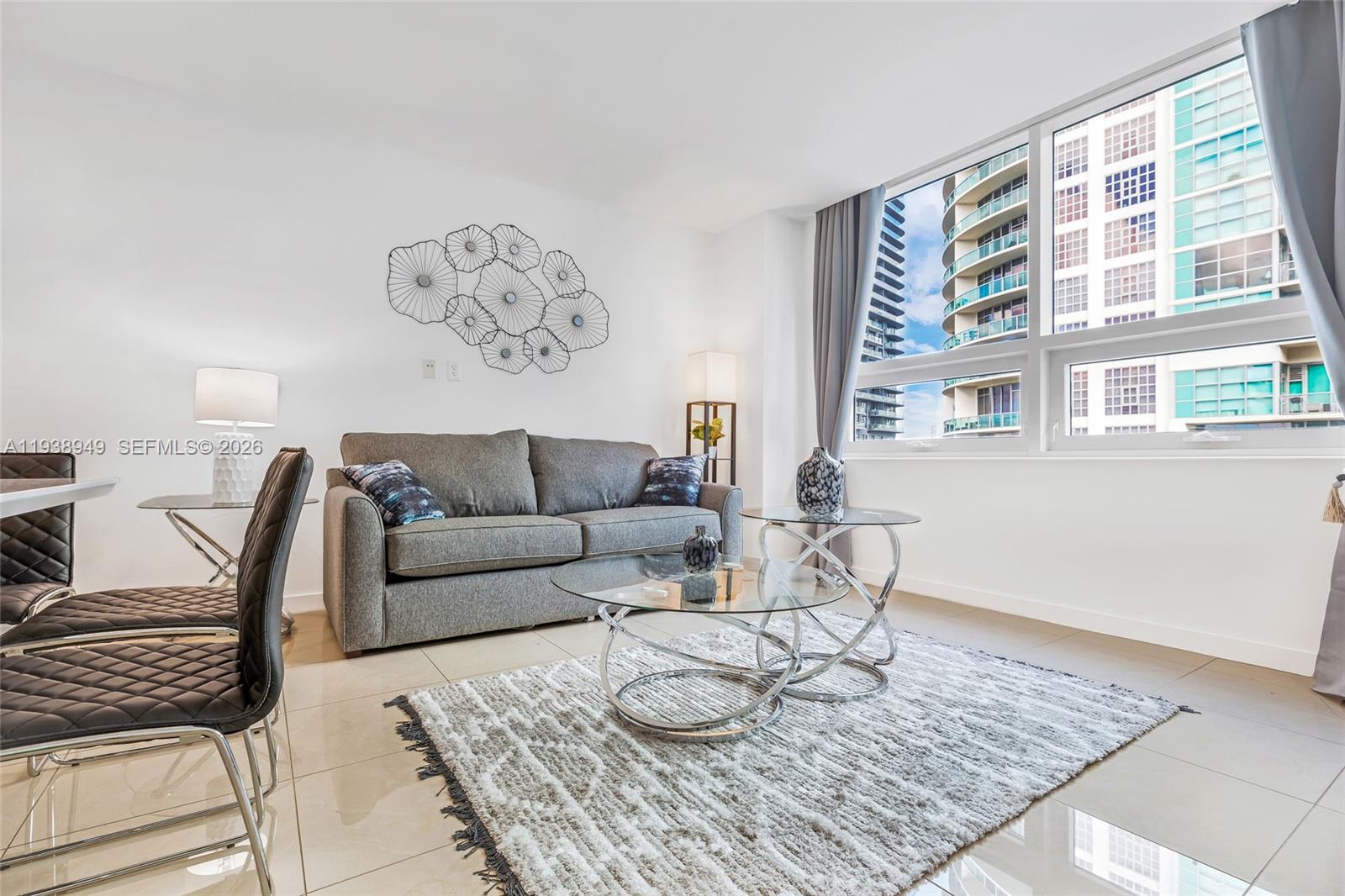 A living room features a grey sofa, glass coffee tables, and a view of buildings — 3250 NE 1st Ave # 603, Miami FL 33137, MIAMI, FL