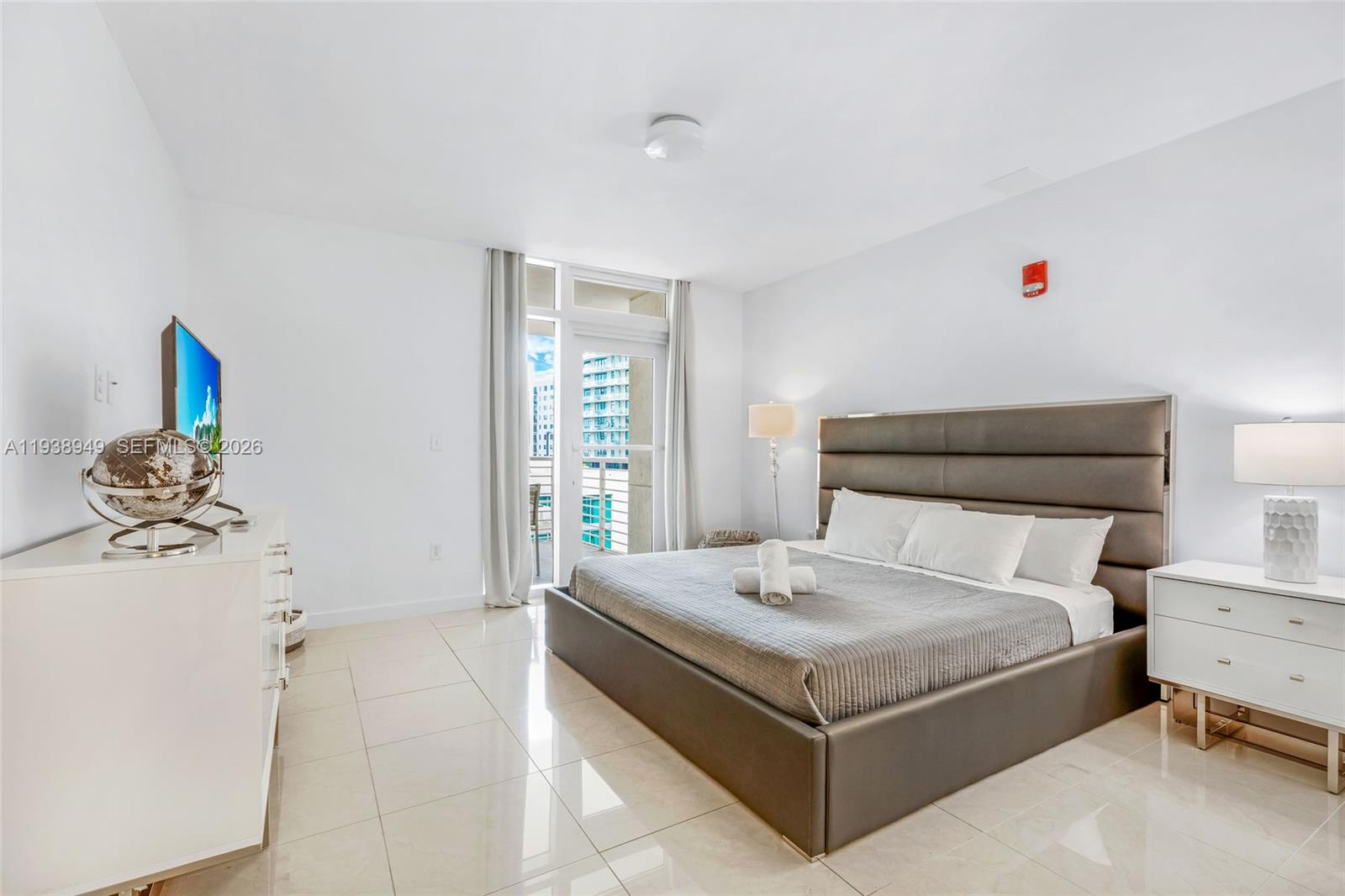 A modern bedroom features a large upholstered bed, white tile floors, and a balcony door — 3250 NE 1st Ave # 603, Miami FL 33137, MIAMI, FL