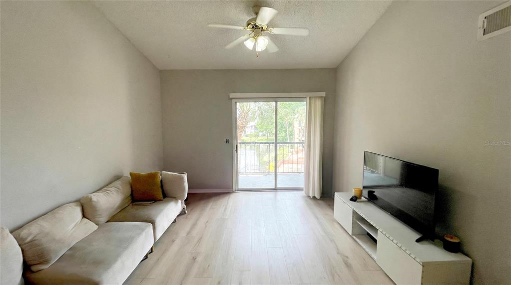 A living room features light wood-look flooring, a beige sofa, a television, and a balcony — 1972 LAKE ATRIUMS CIRCLE UNIT 200, ORLANDO, FL