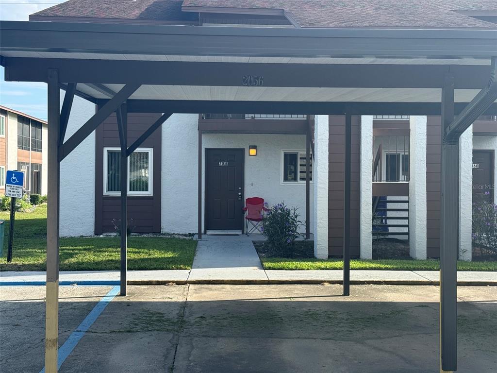 A ground-level apartment entrance features a dark door, a red chair, and a metal carport — 39132 COUNTY RD. 54 UNIT 2056, ZEPHYRHILLS, FL