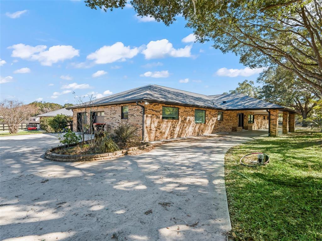 A brick ranch-style house features a concrete driveway, a covered carport, and mature trees — 9865 SW 74TH AVENUE, OCALA, FL