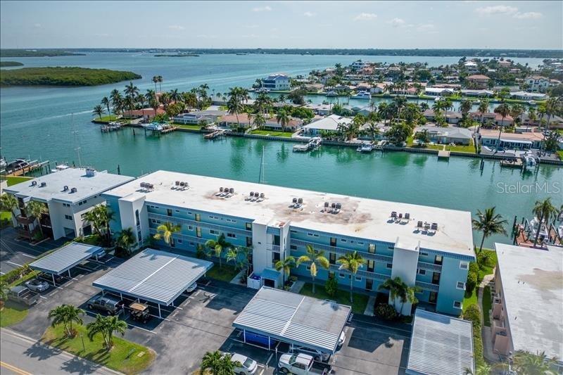 An aerial view shows a multi-story blue apartment building with covered parking and waterfront access — 12405 3RD STREET E UNIT 201, TREASURE ISLAND, FL
