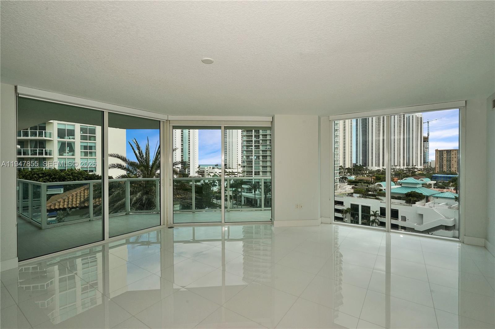 An empty room features white tile flooring, floor-to-ceiling windows, and a wrap-around balcony — SUNNY ISLES BEACH, FL