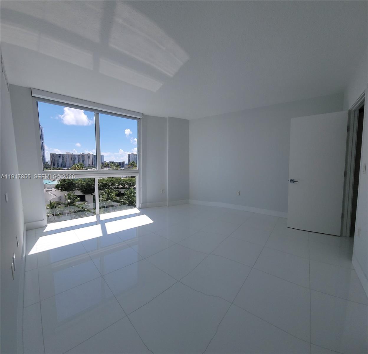 An empty room features white tile floors, white walls, and a large window with city views — SUNNY ISLES BEACH, FL