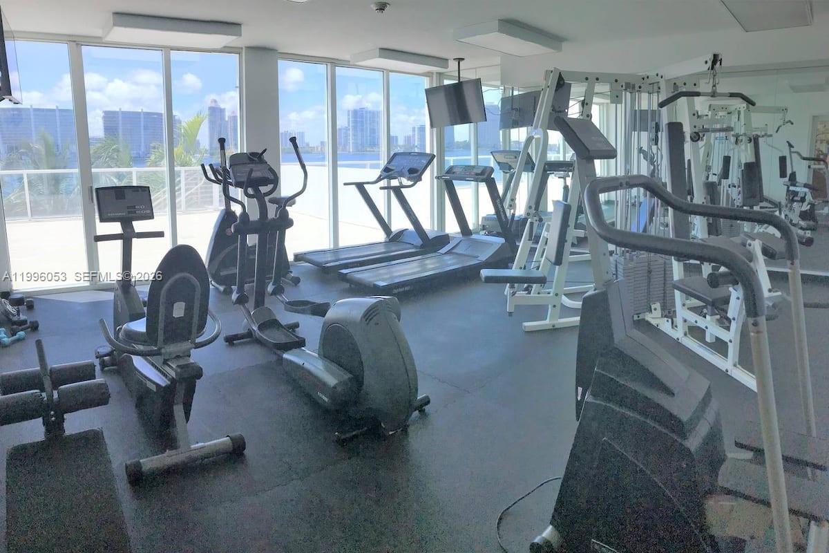 This gym features various exercise machines and floor-to-ceiling windows overlooking a waterfront city skyline — SUNNY ISLES BEACH, FL