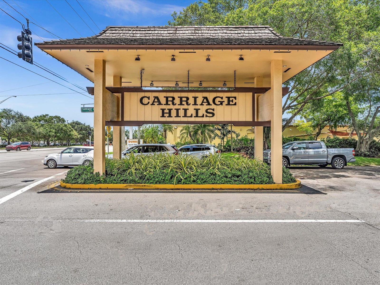 A large wooden entrance sign for Carriage Hills stands over a landscaped island with cars — 100 Ashbury Road # 103, Hollywood FL 33024, HOLLYWOOD, FL