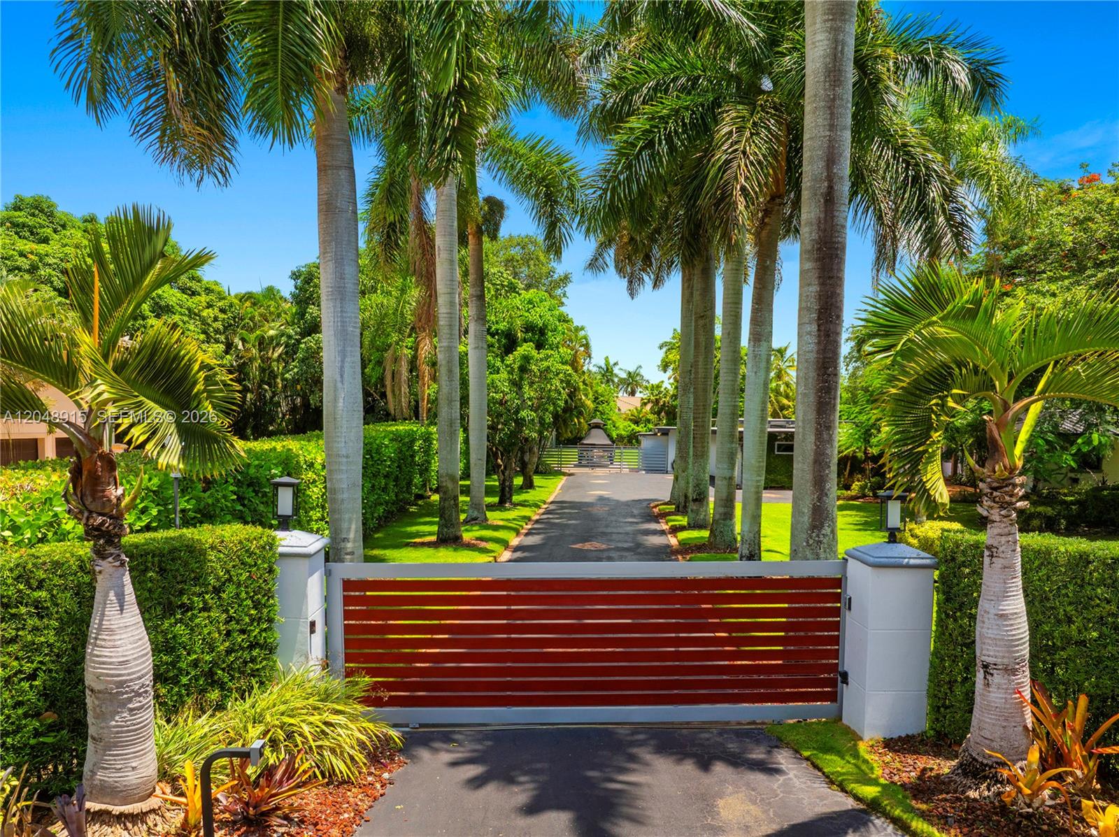 A gated driveway leads past tall palm trees toward a house on a sunny day — 8735 SW 122nd St, Miami FL 33176, MIAMI, FL