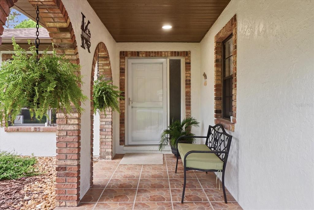 A covered front porch features brick arches, tiled flooring, a metal bench, and hanging plants — 5 DAHOON DRIVE, HOMOSASSA, FL