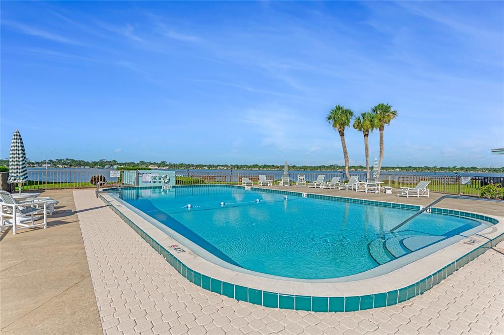 An outdoor swimming pool with a tiled edge overlooks a lake with palm trees nearby — 3009 N HALIFAX AVENUE UNIT 1, DAYTONA BEACH, FL