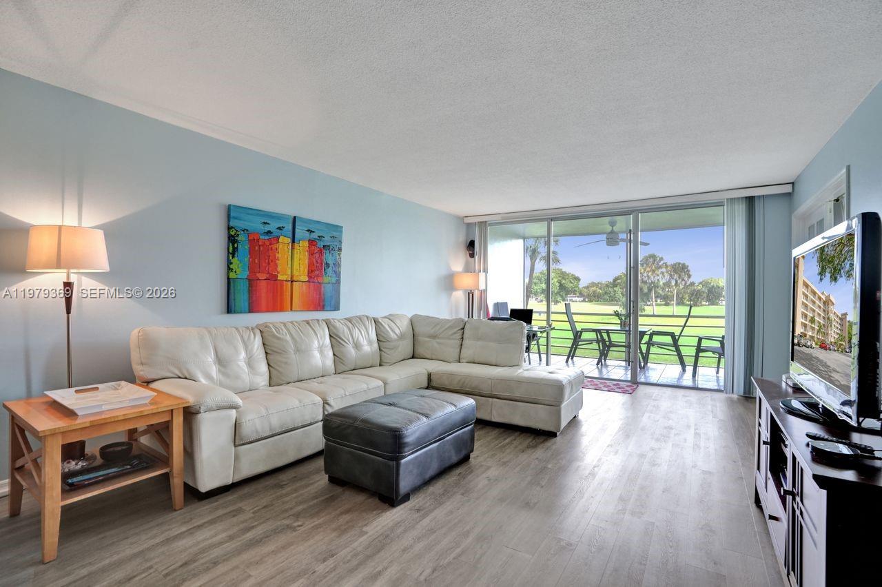 A living room features a white sectional sofa, gray ottoman, and sliding doors to a balcony — 2651 S Course Dr # 203, Pompano Beach FL 33069, POMPANO BEACH, FL