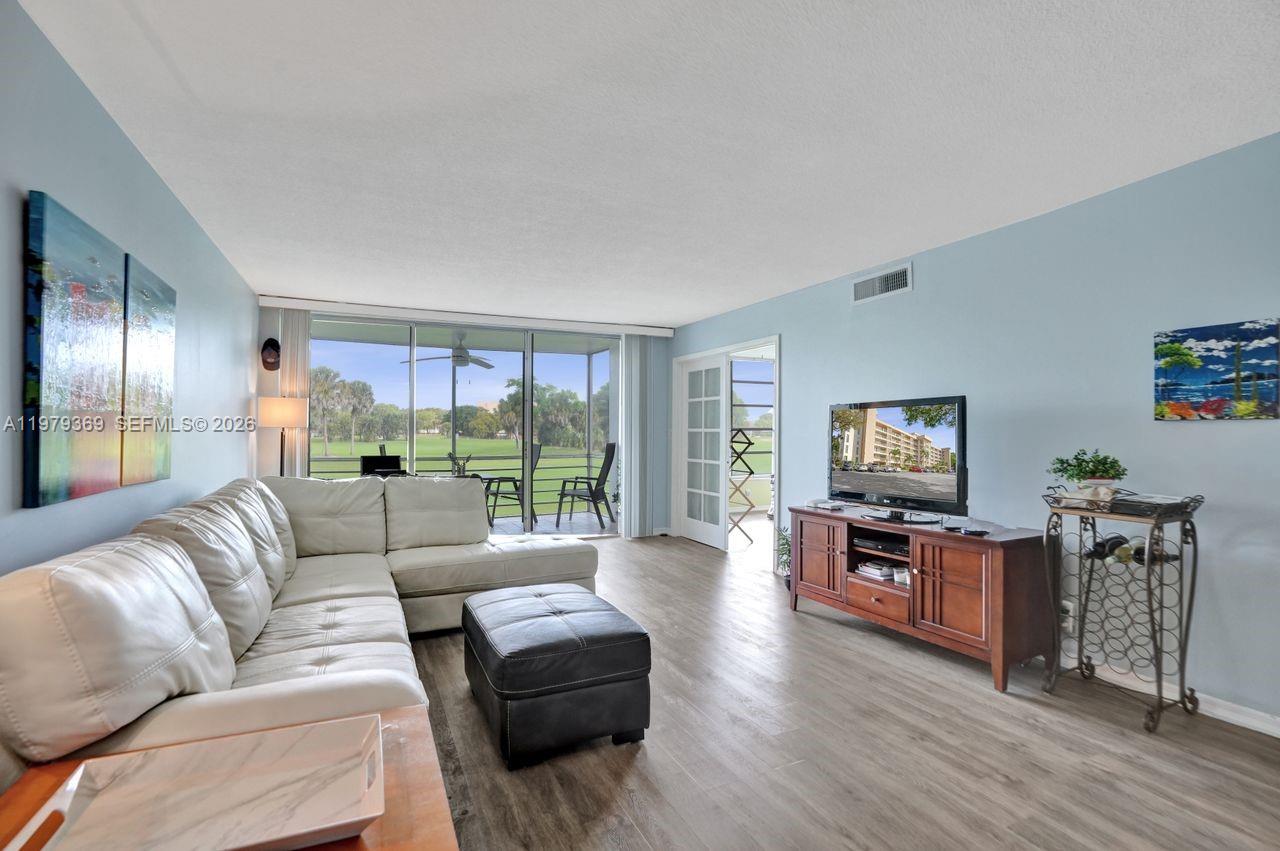 A living room features a white sectional sofa, wood-look flooring, and a balcony with golf views — 2651 S Course Dr # 203, Pompano Beach FL 33069, POMPANO BEACH, FL