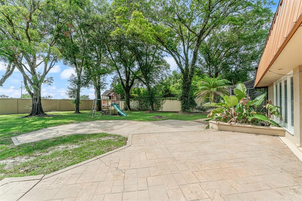 A backyard patio features stone pavers, a wooden playset, and mature trees against a fence — 229 FLAME AVENUE, MAITLAND, FL