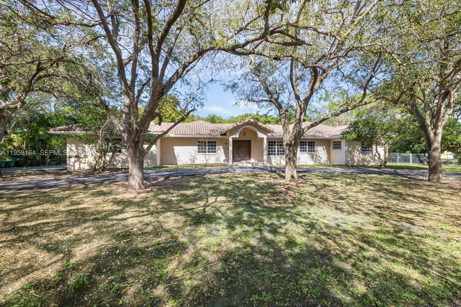 A single-story house with a tile roof and a circular driveway sits behind large trees — 7821 SW 129th Ter, Pinecrest FL 33156, PINECREST, FL