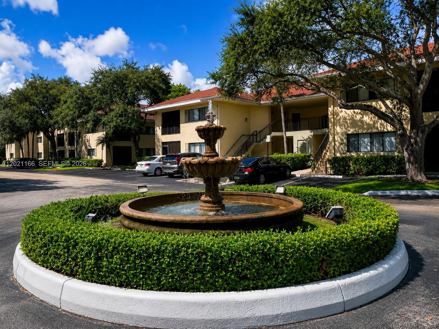 A tiered stone fountain sits in a circular hedge garden in front of apartment buildings — FORT LAUDERDALE, FL