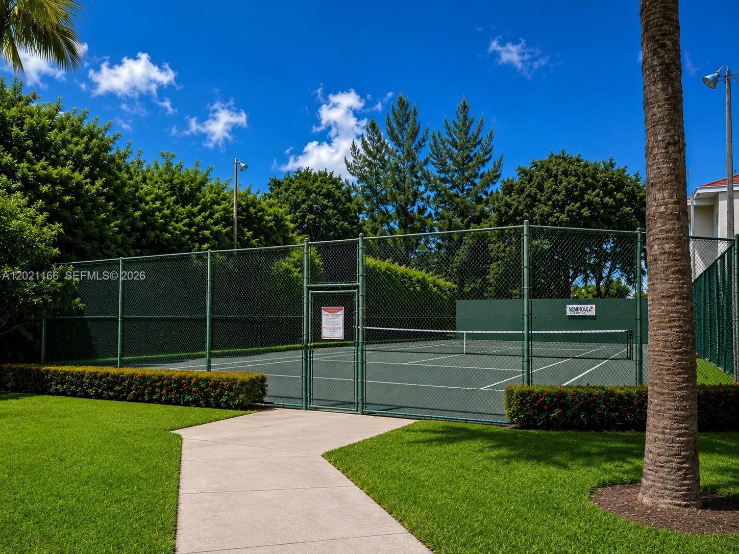 A fenced tennis court sits outdoors next to a paved walkway and green lawn — FORT LAUDERDALE, FL