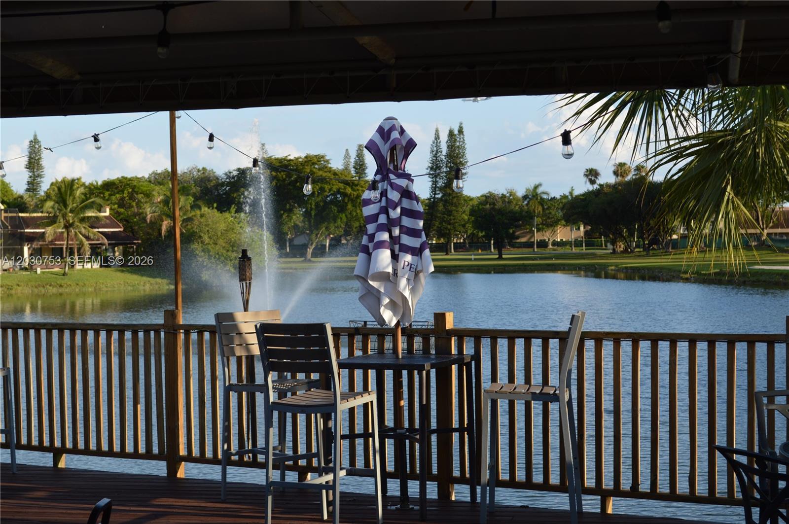 A wooden deck overlooks a lake with a fountain, featuring outdoor seating and string lights — 3932 Estepona Ave, Doral FL 33178, DORAL, FL