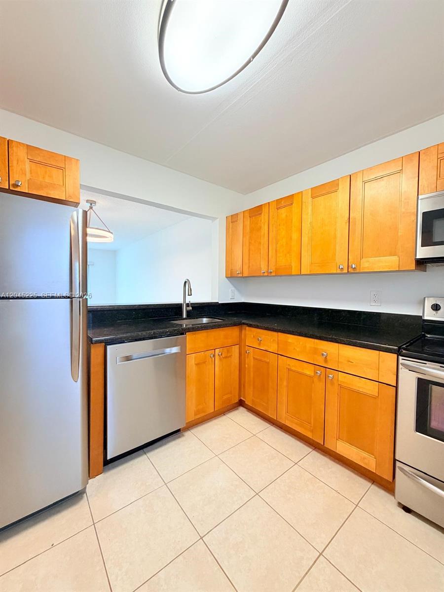 A kitchen features light wood cabinets, black countertops, stainless steel appliances, and tile flooring — 7705 Camino Real # B413, Miami FL 33143, MIAMI, FL